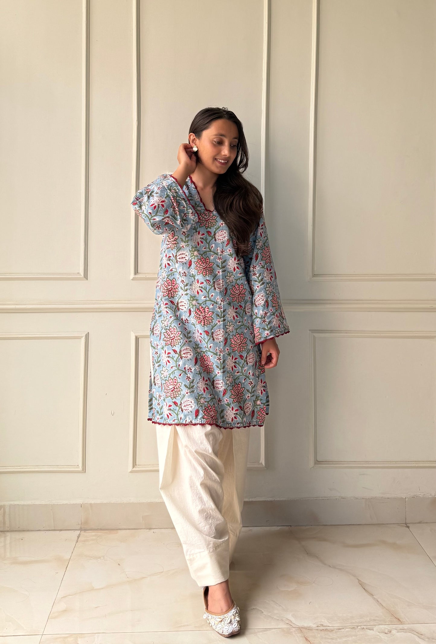 Woman wearing a floral top and white pants against a light-colored paneled wall.