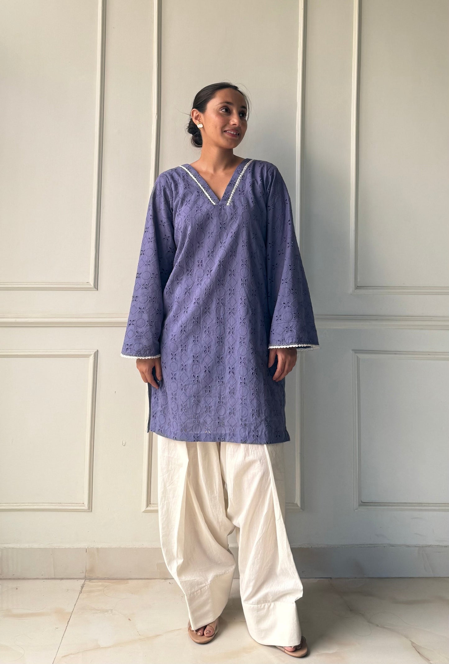 Woman wearing a blue kurta with white pants against a white paneled wall.