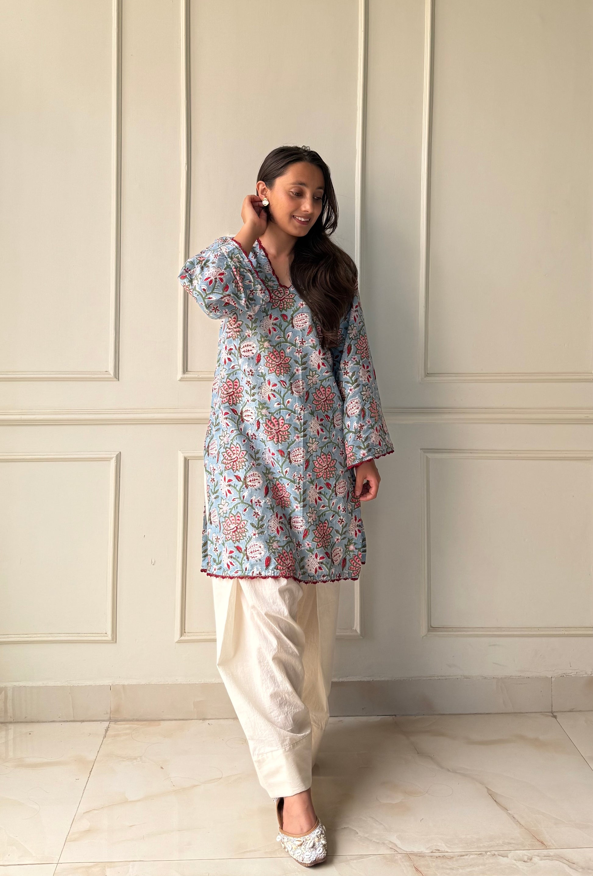 Woman wearing a floral top and white pants against a light-colored paneled wall.