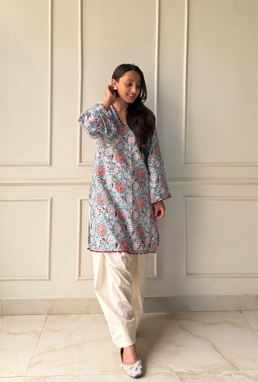 Woman wearing a floral top and white pants against a light-colored paneled wall.