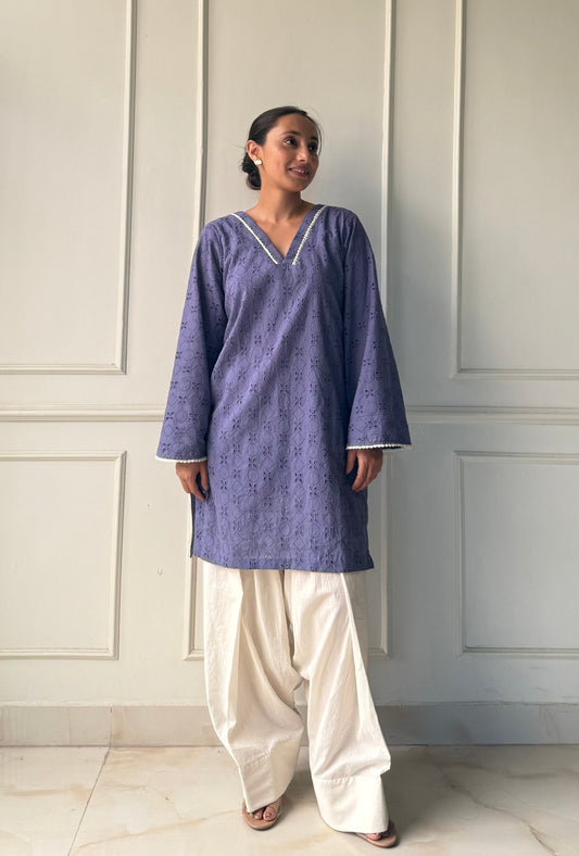 Woman wearing a blue kurta with white pants against a white paneled wall.